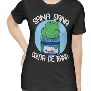 May include: Dark gray t-shirt featuring a cartoon frog sitting on a jar of VapoR&uacute;. The frog is green with a bandage, and the text "Sana Sana Colita de Rana" is written around the image in white.