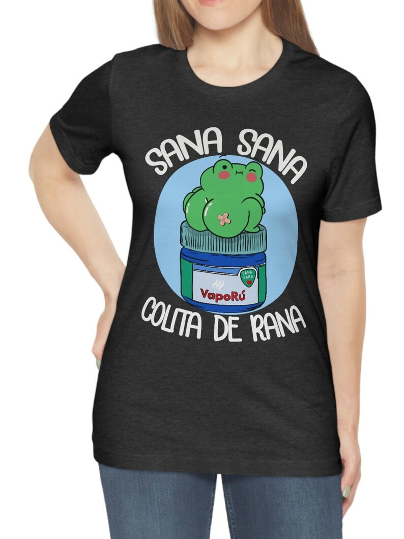 May include: Dark gray t-shirt featuring a cartoon frog sitting on a jar of VapoR&uacute;. The frog is green with a bandage, and the text "Sana Sana Colita de Rana" is written around the image in white.