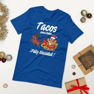 May include: Royal blue t-shirt with a festive graphic. The design features a luchador in a Santa hat riding a sleigh pulled by reindeer, with the text "Tacos para todos... &iexcl;Feliz Navidad!"