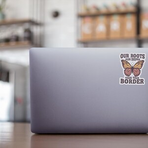 K&ouml;nnte beinhalten: Ein grauer Laptop mit einem Aufkleber mit einem Schmetterlingsdesign und dem Text "OUR ROOTS RUN DEEPER THAN YOUR BORDER". Der Schmetterling hat orange und rosa Fl&uuml;gel.
