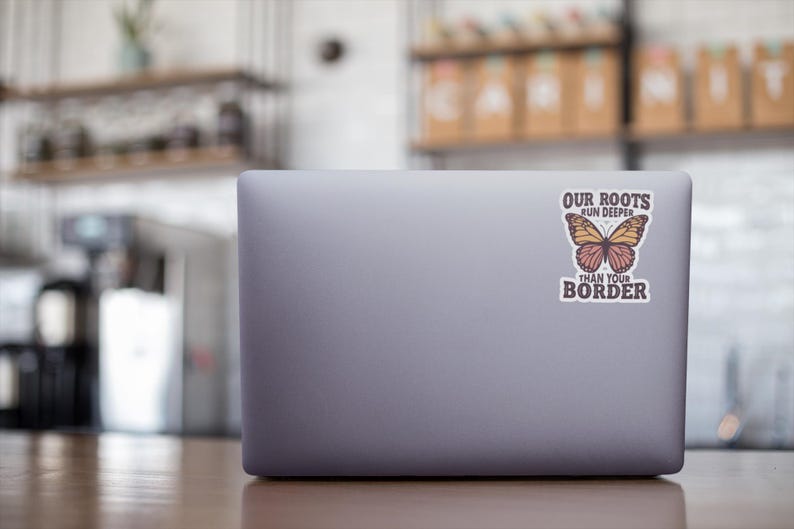 K&ouml;nnte beinhalten: Ein grauer Laptop mit einem Aufkleber mit einem Schmetterlingsdesign und dem Text "OUR ROOTS RUN DEEPER THAN YOUR BORDER". Der Schmetterling hat orange und rosa Fl&uuml;gel.