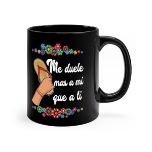 May include: Black coffee mug with a white graphic of a flip-flop and the text "Me duele mas a mi que a ti" surrounded by colorful flowers.