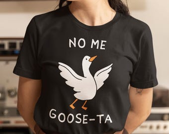 May include: Black t-shirt with the text "NO ME GOOSE-TA" and a white goose graphic with orange beak and feet. The goose has outstretched wings. The shirt is worn by a person.