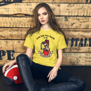 May include: A yellow t-shirt featuring a graphic of a skeleton woman flexing her bicep with a rose in her hair. The text on the shirt reads "And she will raise to be Chingona".