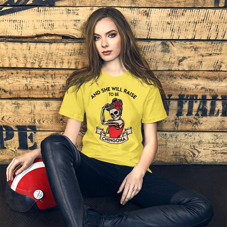 May include: A yellow t-shirt featuring a graphic of a skeleton woman flexing her bicep with a rose in her hair. The text on the shirt reads "And she will raise to be Chingona".