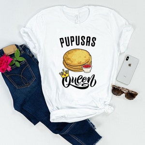 Puede incluir: Camiseta blanca con texto negro que dice "Pupusas Queen" y un gr&aacute;fico de una pila de pupusas con un peque&ntilde;o taz&oacute;n de salsa roja y una corona dorada.