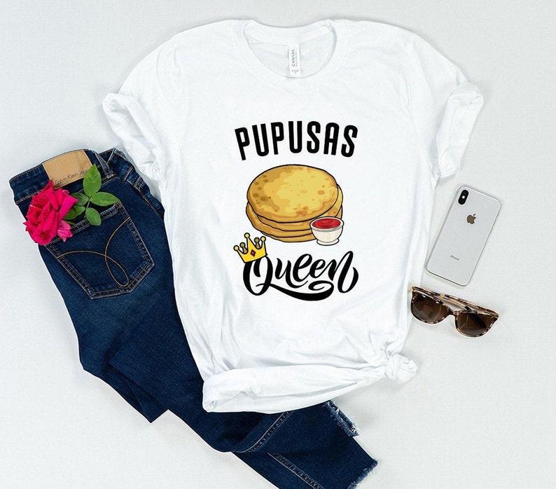 Puede incluir: Camiseta blanca con texto negro que dice "Pupusas Queen" y un gr&aacute;fico de una pila de pupusas con un peque&ntilde;o taz&oacute;n de salsa roja y una corona dorada.