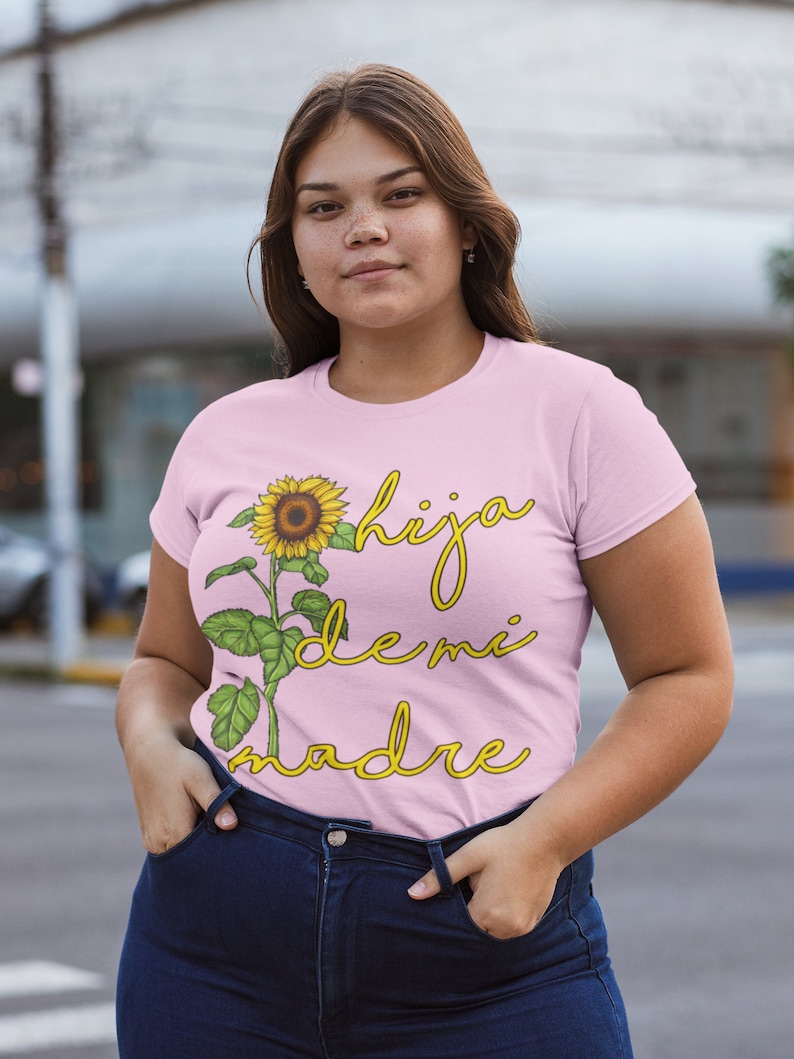 Puede incluir: Una camiseta rosa con un gr&aacute;fico de girasol y el texto "hija de mi madre" en letra amarilla.