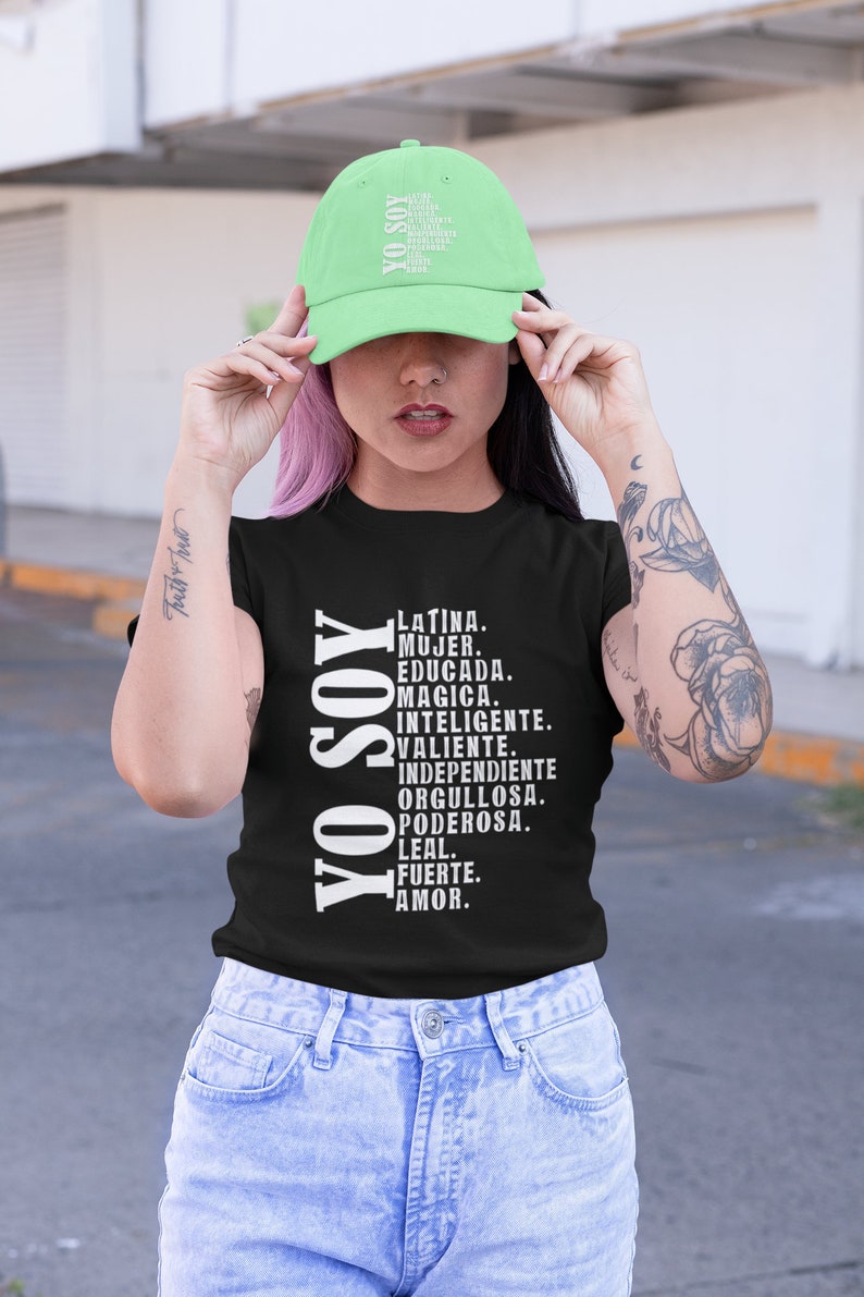 Puede incluir: Una camiseta negra con el texto "YO SOY" en blanco, seguido de una lista de palabras en espa&ntilde;ol que describen las cualidades de una mujer. La modelo lleva una gorra de b&eacute;isbol verde claro con el mismo texto en blanco.