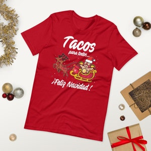 May include: Red t-shirt with a festive graphic. The design features a taco character in a Santa hat riding a sleigh pulled by reindeer, with the text "Tacos para todos... &iexcl;Feliz Navidad!"