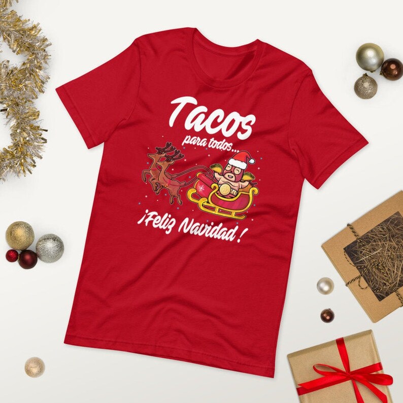 May include: Red t-shirt with a festive graphic. The design features a taco character in a Santa hat riding a sleigh pulled by reindeer, with the text "Tacos para todos... &iexcl;Feliz Navidad!"