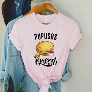 Puede incluir: Camiseta rosa con el texto "Pupusas Queen" en letras negras. Un gr&aacute;fico de una pila de pupusas con una corona y un peque&ntilde;o taz&oacute;n de salsa roja est&aacute; por encima del texto.