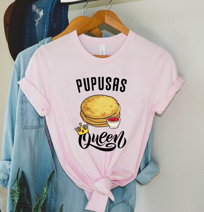 Puede incluir: Camiseta rosa con el texto "Pupusas Queen" en letras negras. Un gr&aacute;fico de una pila de pupusas con una corona y un peque&ntilde;o taz&oacute;n de salsa roja est&aacute; por encima del texto.