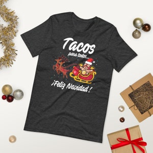 May include: Dark gray t-shirt with a festive design. The shirt features the text "Tacos para todos... &iexcl;Feliz Navidad!" above an illustration of a taco in a Santa hat riding a sleigh pulled by reindeer. A fun, holiday-themed tee.