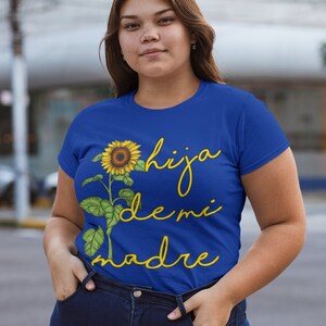 Puede incluir: Una camiseta azul real con un gr&aacute;fico de girasol amarillo y el texto "hija de mi madre" en amarillo.