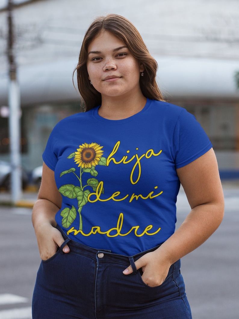 Puede incluir: Una camiseta azul real con un gr&aacute;fico de girasol amarillo y el texto "hija de mi madre" en amarillo.