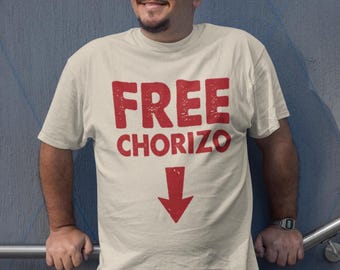 Könnte beinhalten: Cremefarbenes T-Shirt mit dem Text "FREE CHORIZO" in verblichenen roten Buchstaben und einem roten Pfeil nach unten. Das Shirt hat kurze Ärmel und scheint aus Baumwolle zu sein.