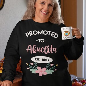 May include: Black sweatshirt with the text "PROMOTED - TO - Abuelita EST. 2019" in white and pink. The design includes a floral and leaf arrangement. A white mug with the text "BUT FIRST Cafecito" is held in the hand.