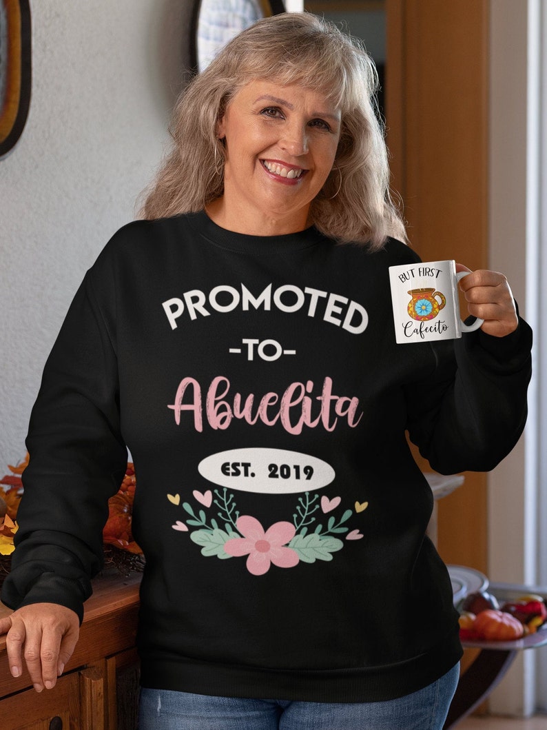 May include: Black sweatshirt with the text "PROMOTED - TO - Abuelita EST. 2019" in white and pink. The design includes a floral and leaf arrangement. A white mug with the text "BUT FIRST Cafecito" is held in the hand.