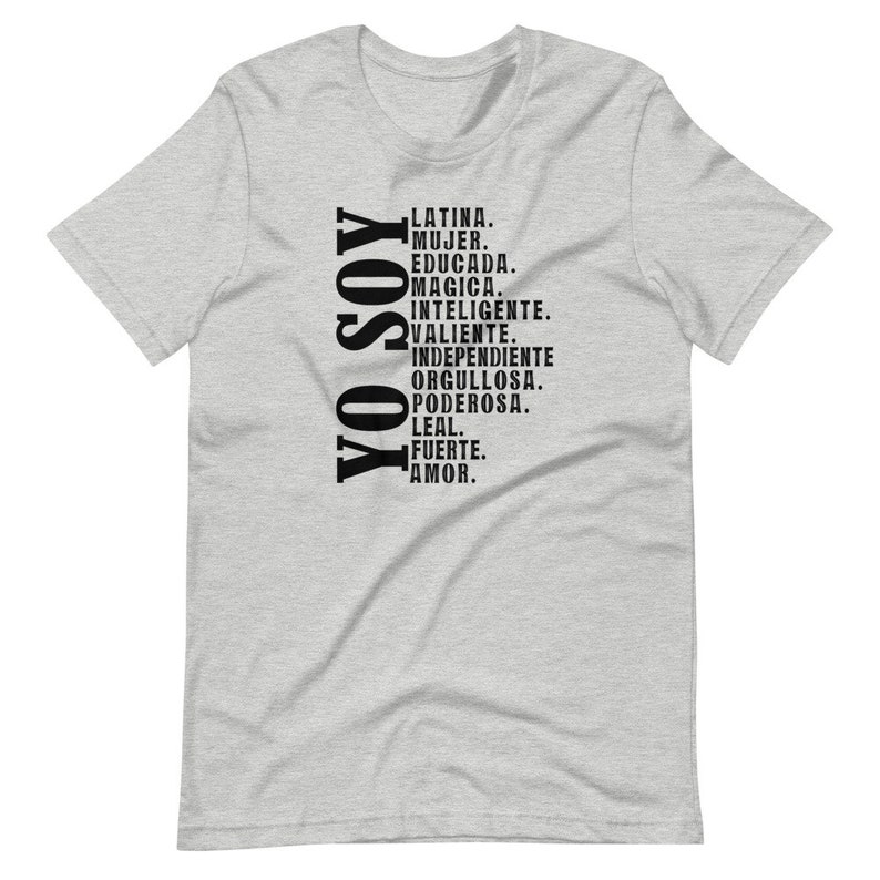 Puede incluir: Una camiseta gris con texto negro que dice "YO SOY" verticalmente, seguida de una lista de palabras en espa&ntilde;ol: "LATINA. MUJER. EDUCADA. MAGICA. INTELIGENTE. VALIENTE. INDEPENDIENTE ORGULLOSA. PODEROSA. LEAL. FUERTE. AMOR."