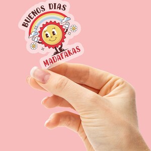 Op de afbeelding: Een sticker met een cartoonzon en regenboog, met de tekst "BUENOS DIAS MADAFAKAS" in een kleurrijk lettertype.