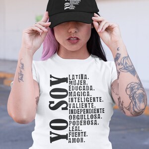 Puede incluir: Una camiseta blanca con el texto "YO SOY" en negro, seguido de una lista de palabras en espa&ntilde;ol que describen a una mujer latina. Las palabras incluyen "Latina, Mujer, Educada, Magica, Inteligente, Valiente, Independiente, Orgullosa, Poderosa, Leal, Fuerte, Amor."