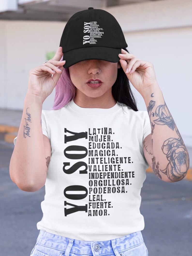 Puede incluir: Una camiseta blanca con el texto "YO SOY" en negro, seguido de una lista de palabras en espa&ntilde;ol que describen a una mujer latina. Las palabras incluyen "Latina, Mujer, Educada, Magica, Inteligente, Valiente, Independiente, Orgullosa, Poderosa, Leal, Fuerte, Amor."
