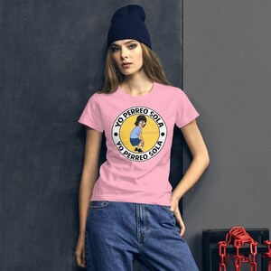 May include: A pink t-shirt with a white circle graphic featuring a cartoon woman with brown hair and glasses. The text "Yo Perreo Sola" is written around the circle in black.