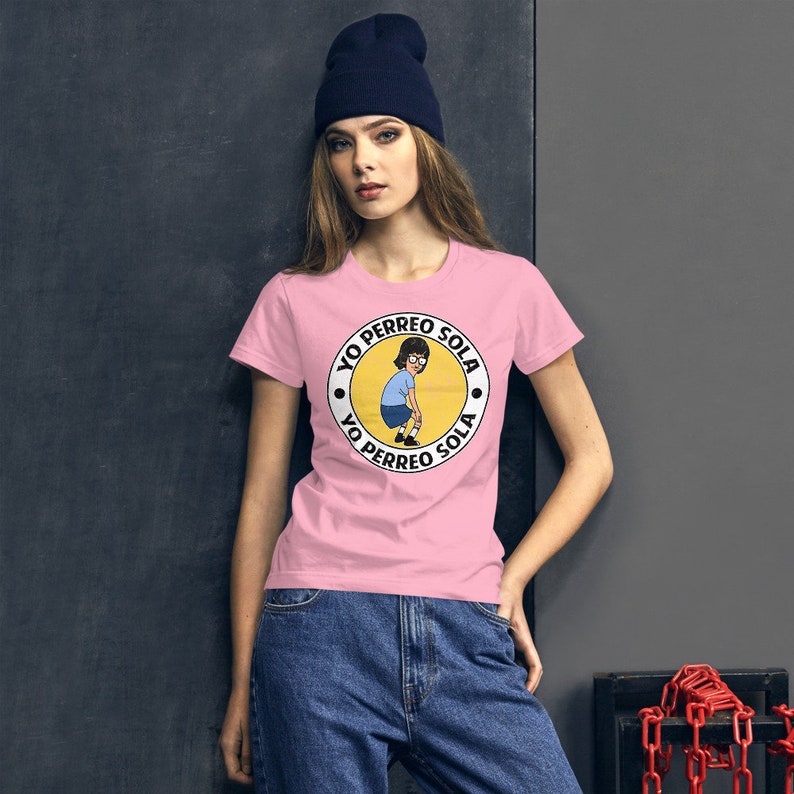 May include: A pink t-shirt with a white circle graphic featuring a cartoon woman with brown hair and glasses. The text "Yo Perreo Sola" is written around the circle in black.