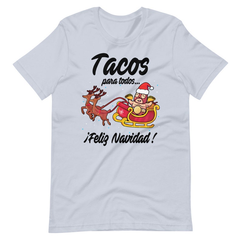 May include: Light blue t-shirt with a festive graphic. The design features a luchador taco in a Santa hat riding a sleigh pulled by reindeer, with the text "Tacos para todos... &iexcl;Feliz Navidad!"