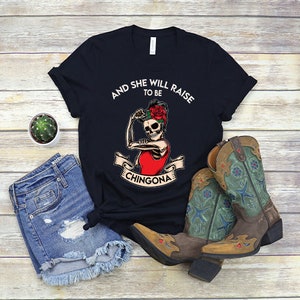 May include: Black t-shirt featuring a white graphic of a skeleton woman with a red flower in her hair. The text "And she will raise to be Chingona" is printed on the shirt.