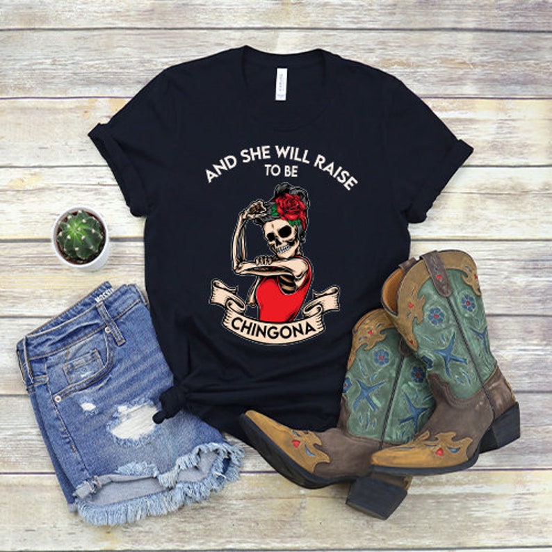 May include: Black t-shirt featuring a white graphic of a skeleton woman with a red flower in her hair. The text "And she will raise to be Chingona" is printed on the shirt.