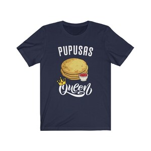 Puede incluir: Una camiseta azul marino con el texto "Pupusas Queen" en letras blancas. Una imagen de dibujos animados de pupusas con un lado de salsa est&aacute; por encima del texto. Una corona dorada est&aacute; a la izquierda del texto.
