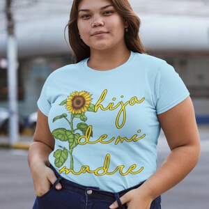 Puede incluir: Una camiseta azul claro con un gr&aacute;fico de girasol y el texto "hija de mi madre" en amarillo.