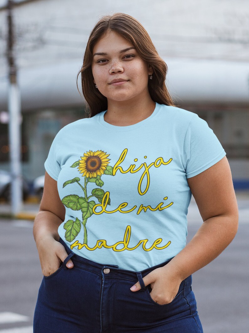 Puede incluir: Una camiseta azul claro con un gr&aacute;fico de girasol y el texto "hija de mi madre" en amarillo.