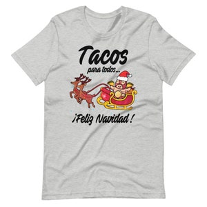 May include: A light gray t-shirt featuring a festive design. The graphic shows a luchador taco in a Santa hat riding a sleigh pulled by reindeer, with the text "Tacos para todos... &iexcl;Feliz Navidad!" in black.