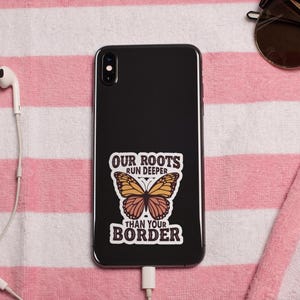 K&ouml;nnte beinhalten: Ein schwarzes Smartphone mit einem Aufkleber mit der Aufschrift "OUR ROOTS RUN DEEPER THAN YOUR BORDER" und einer Schmetterlingsgrafik. Das Telefon liegt auf einem rosa-wei&szlig; gestreiften Handtuch, mit Sonnenbrille und wei&szlig;en Ohrh&ouml;rern in der N&auml;he.
