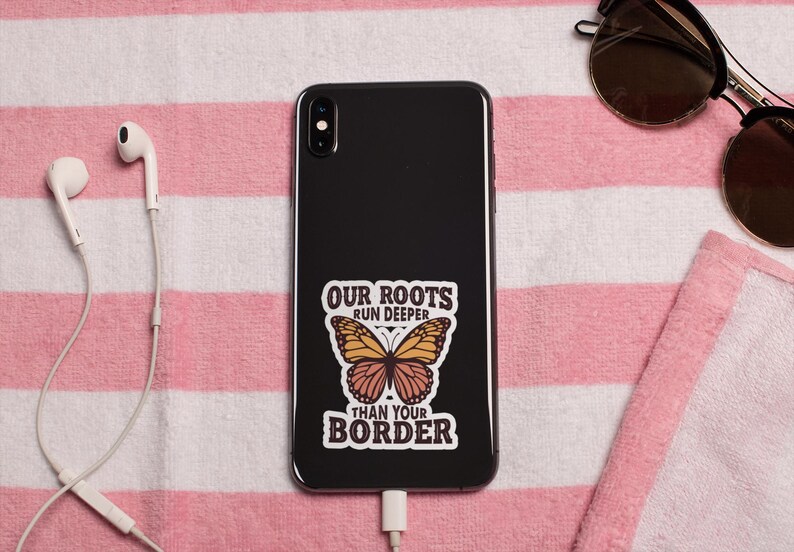 K&ouml;nnte beinhalten: Ein schwarzes Smartphone mit einem Aufkleber mit der Aufschrift "OUR ROOTS RUN DEEPER THAN YOUR BORDER" und einer Schmetterlingsgrafik. Das Telefon liegt auf einem rosa-wei&szlig; gestreiften Handtuch, mit Sonnenbrille und wei&szlig;en Ohrh&ouml;rern in der N&auml;he.