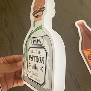 May include: White card shaped like a Patron tequila bottle with the text "Pap&aacute; Feliz D&iacute;a Patr&oacute;n 100% Ching&oacute;n 100% Trabajador Respeto y Admiraci&oacute;n"