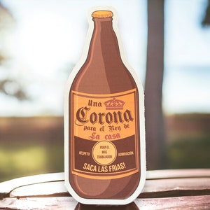 May include: A brown beer bottle with a white label that says "Una Corona para el Rey de la casa". The label also has the words "Respeto", "Para el Mas Trabajador", and "Admiraci&oacute;n". The bottle is sitting on a wooden surface.