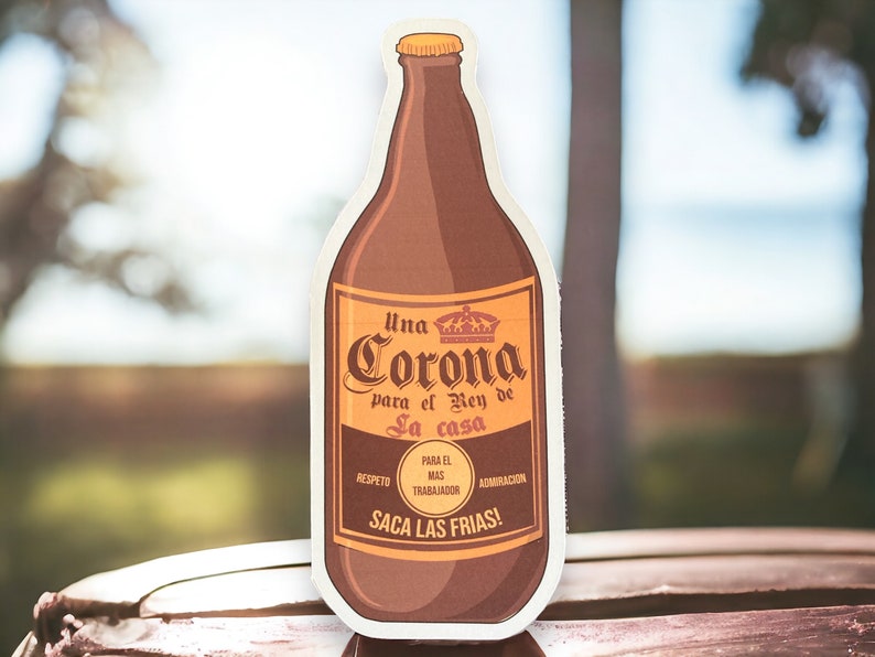 May include: A brown beer bottle with a white label that says "Una Corona para el Rey de la casa". The label also has the words "Respeto", "Para el Mas Trabajador", and "Admiraci&oacute;n". The bottle is sitting on a wooden surface.