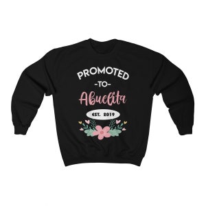 May include: Black sweatshirt with the text "PROMOTED -to- Abuelita EST. 2019" in white and pink lettering. The design includes a floral arrangement with pink flowers and green leaves.