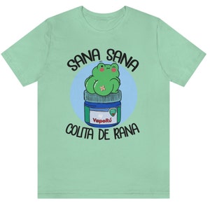 May include: A light green t-shirt featuring a cartoon frog design. The frog is green and sitting on a blue and white jar. The text "SANA SANA COLITA DE RANA" is printed above and below the frog.