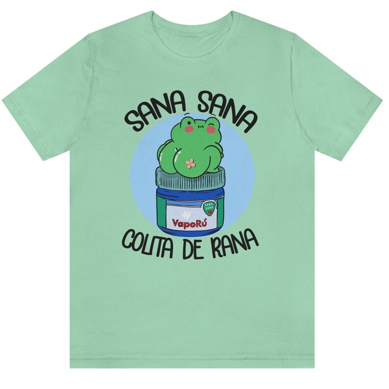May include: A light green t-shirt featuring a cartoon frog design. The frog is green and sitting on a blue and white jar. The text "SANA SANA COLITA DE RANA" is printed above and below the frog.