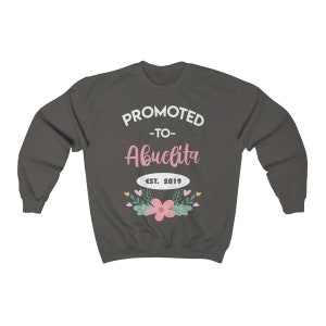 May include: Dark gray sweatshirt with the text "PROMOTED -to- Abuelita EST. 2019" in white and pink. The design includes a floral arrangement with pink flowers and green leaves. A cozy, casual garment.