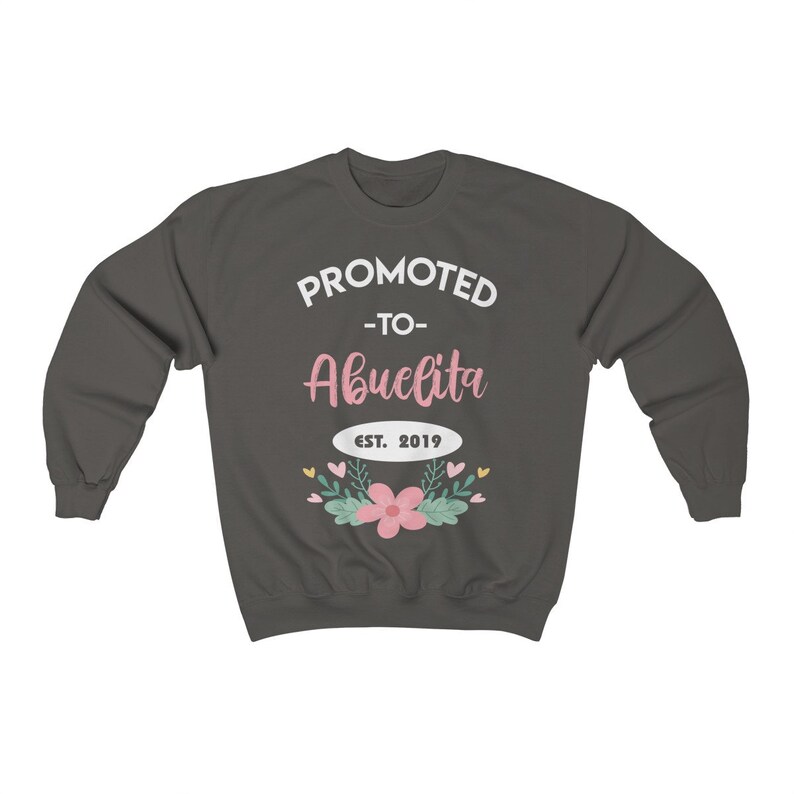 May include: Dark gray sweatshirt with the text "PROMOTED -to- Abuelita EST. 2019" in white and pink. The design includes a floral arrangement with pink flowers and green leaves. A cozy, casual garment.