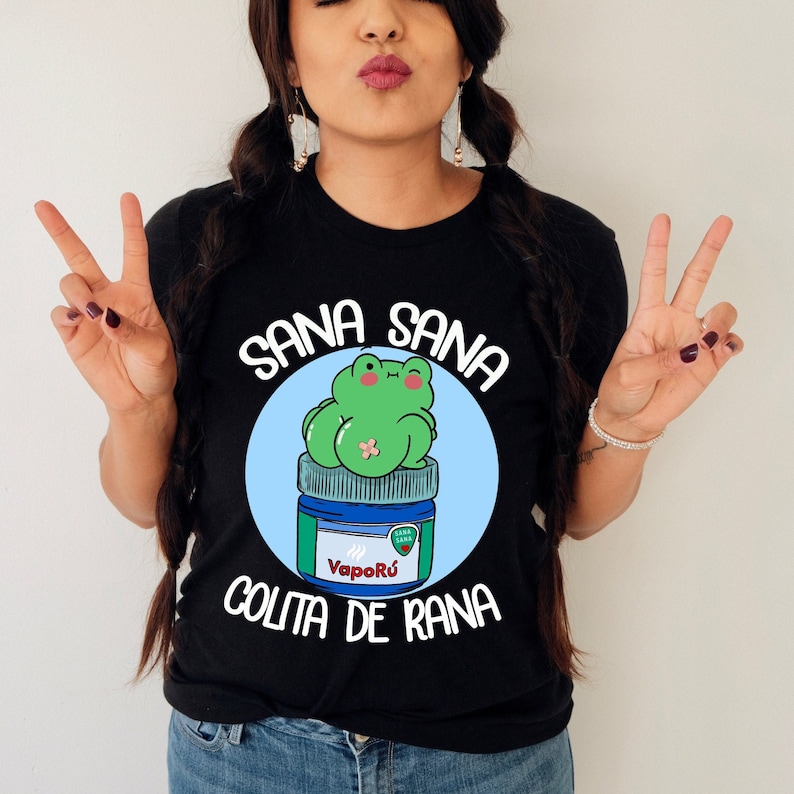 May include: Black t-shirt with a graphic featuring a green frog sitting on a blue and white jar of VapoRub. The text "Sana Sana" is arched above the frog, and "Colita de Rana" is below. The frog has a bandage and is winking.