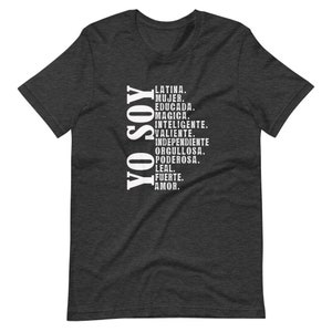 Puede incluir: Una camiseta gris oscuro jaspeada con el texto "YO SOY" en blanco, seguida de una lista de palabras en espa&ntilde;ol que describen las cualidades de una mujer, incluyendo "Latina", "Mujer", "Educada", "Magica", "Inteligente", "Valiente", "Independiente", "Orgullosa", "Poderosa", "Leal", "Fuerte" y "Amor."
