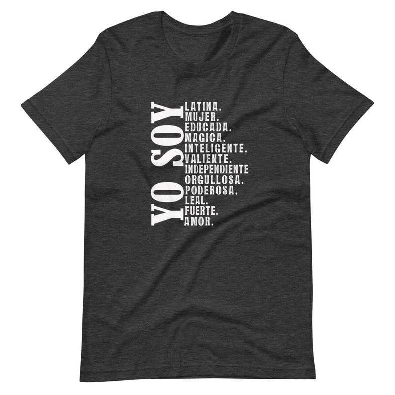 Puede incluir: Una camiseta gris oscuro jaspeada con el texto "YO SOY" en blanco, seguida de una lista de palabras en espa&ntilde;ol que describen las cualidades de una mujer, incluyendo "Latina", "Mujer", "Educada", "Magica", "Inteligente", "Valiente", "Independiente", "Orgullosa", "Poderosa", "Leal", "Fuerte" y "Amor."