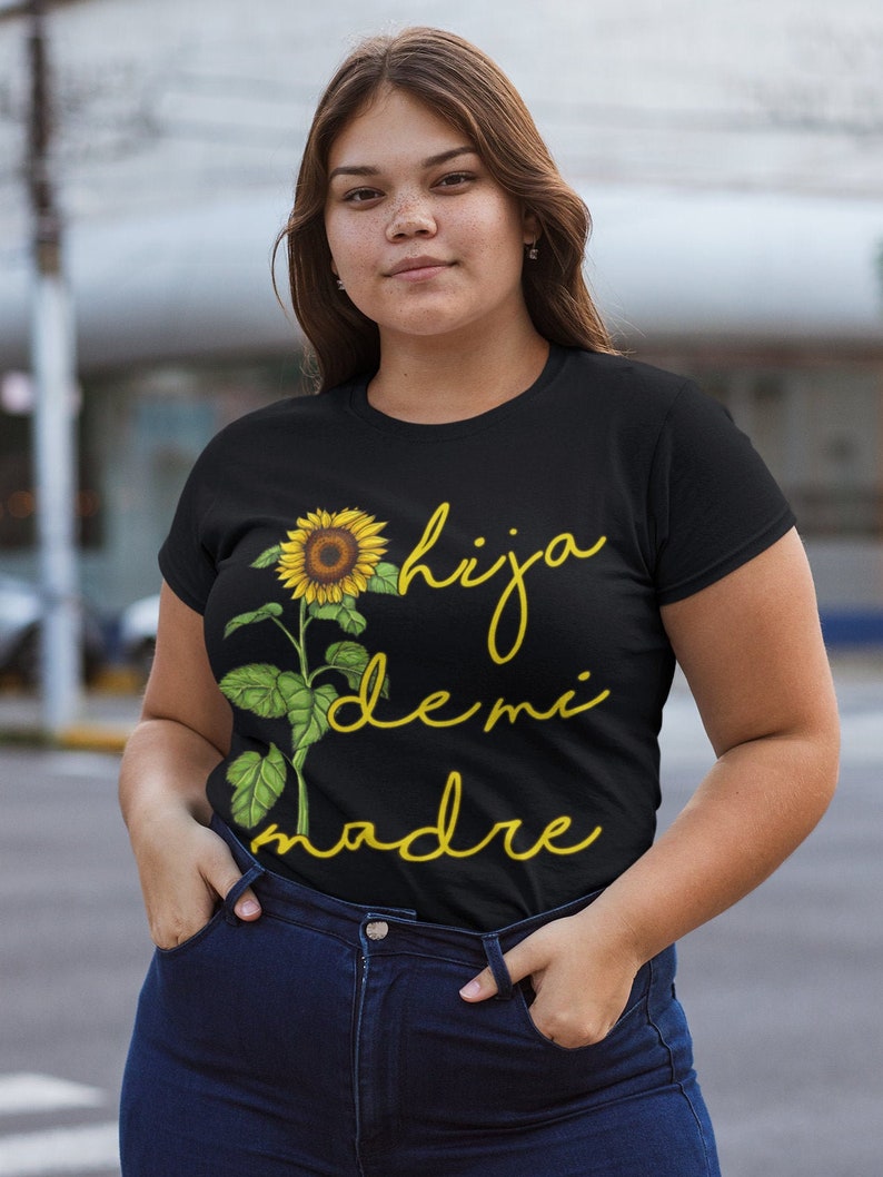 Puede incluir: Camiseta negra con un gr&aacute;fico de girasol y el texto "hija de mi madre" en escritura amarilla.